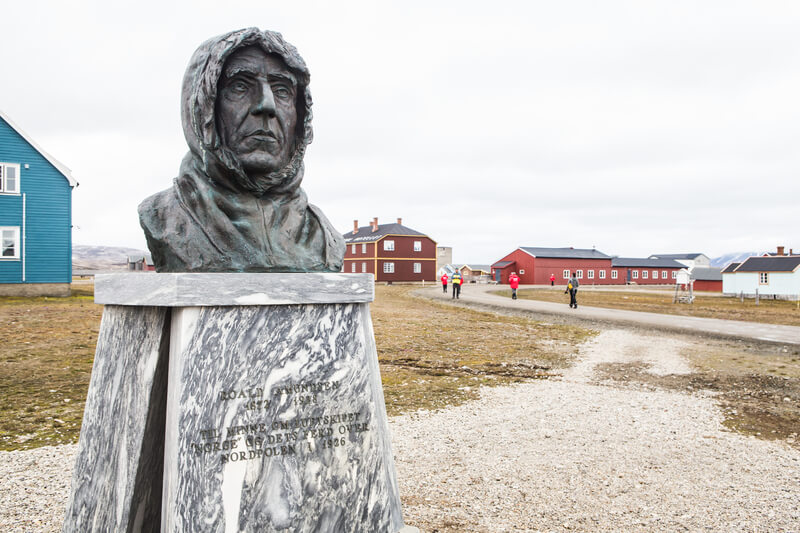 Büste von Roald Amundsen in der kleinen Forschersiedlung Ny-Ålesund mit Kreuzfahrt Gästen die durch den Ort laufen.